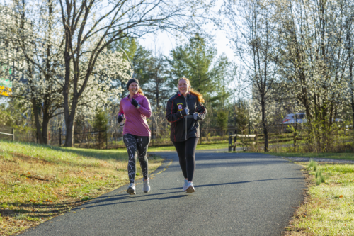 Two smiling women in athletic wear jogging side-by-side along a paved park path lined with blooming white spring trees on a sunny day.